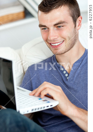 Joyful young man sitting on the sofa using his laptop 6227955