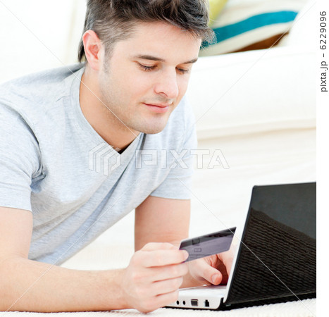 Positive young man holding a card using his laptop on the floor 6229096