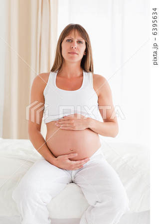Cute pregnant female caressing her belly while sitting on a bed Cute pregnant female caressing her belly while sitting on a bed 6395634