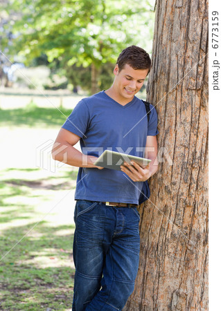 Male student leaning against a tree while using a touch pad Male student leaning against a tree while using a touch pad 6773159