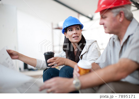 Male and female construction workers sitting down talking and holding coffee and a blueprint 7895171