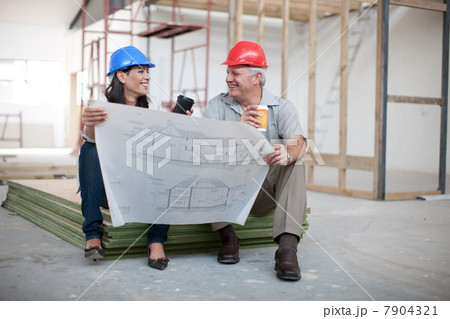 Male and female construction workers sitting down holding coffee and a blueprint 7904321