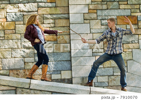 Couple play fighting with sticks against stone wall 7910070