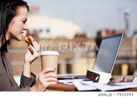 Young woman using a laptop outside while eating a sandwich 7910341