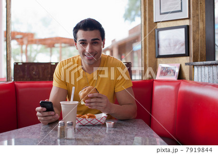 Young man texting on mobile phone and eating fast food in diner, portrait Young man texting on mobile phone and eating fast food in diner, portrait 7919844
