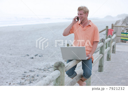 Man sitting on beach railing using phone and laptop 7921232