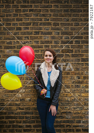 Young woman holding colourful balloons against brick wall 7922167