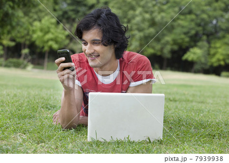 Young man reading text message with laptop in front lying on grass 7939938