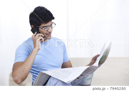 Young man reading newspaper while talking on mobile phone 7940280