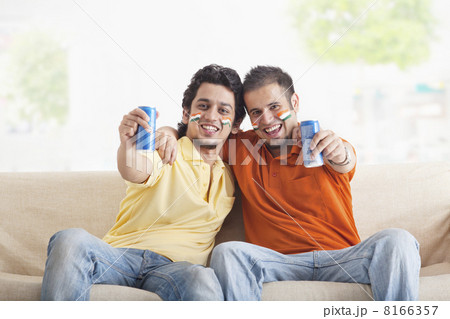 Portrait of cheerful young male friends with face painted holding out tin cans while sitting on sofa 8166357