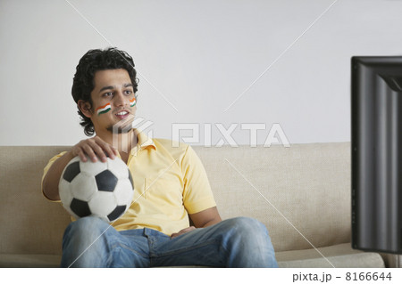 Young man in casuals with face painted watching television while holding soccer ball Young man in casuals with face painted watching television while holding soccer ball 8166644