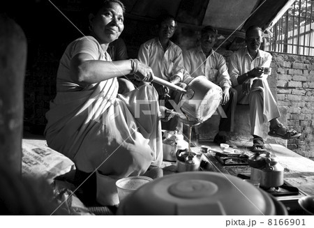 Black and white image of Indian female vendor pouring tea in kettle with customers sitting in background 8166901