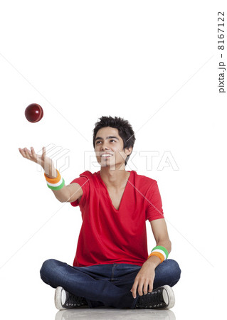 Young boy in casual wear tossing cricket ball while sitting cross-legged over white background 8167122