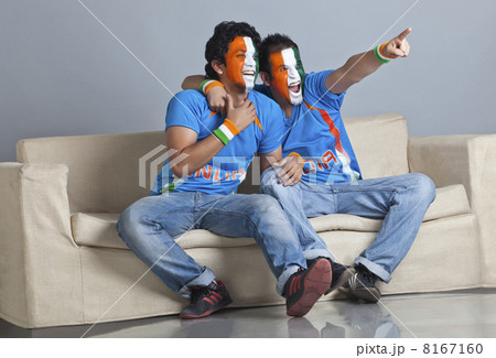 Full length of excited male friends with face painted in Indian tricolor sitting on sofa 8167160