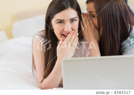 Happy girl telling a secret to her friend in front of laptop Happy girl telling a secret to her friend in front of laptop 8383317
