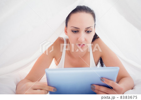 Happy brunette lying under the sheets using her tablet pc Happy brunette lying under the sheets using her tablet pc 8383675