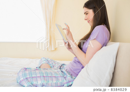 Smiling girl sitting on a bed looking and using a tablet pc 8384029