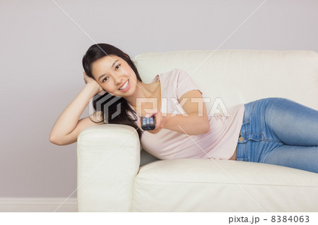 Cheerful asian girl lying on the couch watching tv Cheerful asian girl lying on the couch watching tv 8384063