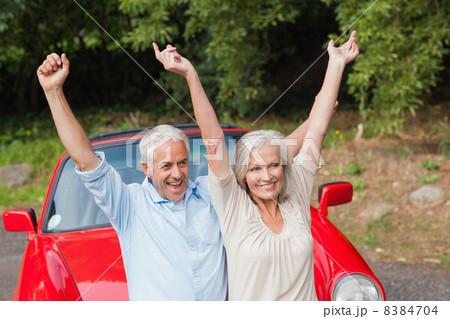 Cheerful mature couple posing by their red convertible Cheerful mature couple posing by their red convertible 8384704