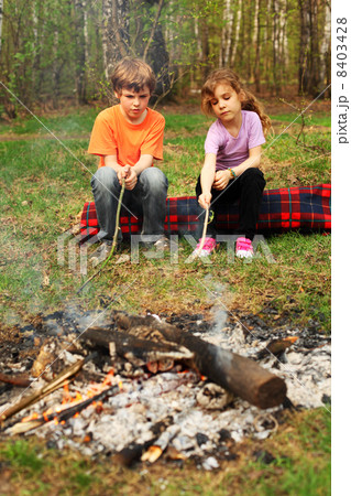 Two children sit around campfire and poke by sticks into ashes 8403428