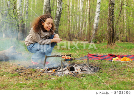 Happy woman sits near campfire with grill and barbecue in forest 8403506