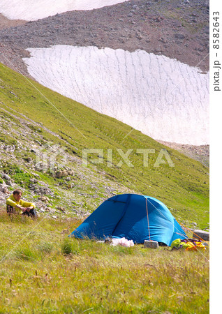 Camping Tent in Mountains with Man Hiker sitting on grass and Glacier Hillside on Background Camping Tent in Mountains with Man Hiker sitting on grass and Glacier Hillside on Background 8582643