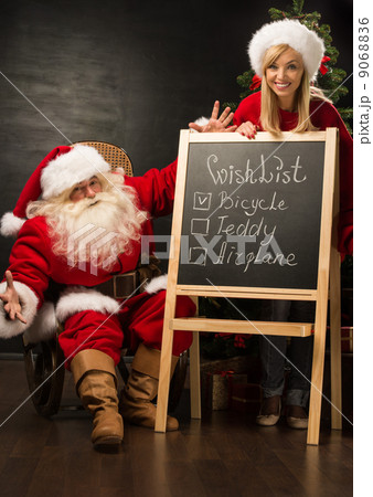 Santa Claus with his helper standing near chalkboard with wishli 9068836