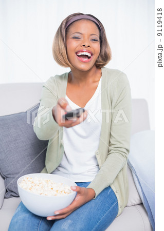 Laughing woman sitting on sofa changing tv channel while holding 9119418