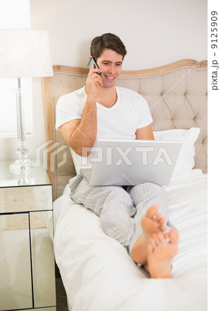 Casual man using cellphone and laptop in bed 9125909