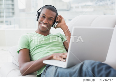 Smiling young Afro man with headphones using laptop on sofa Smiling young Afro man with headphones using laptop on sofa 9128247