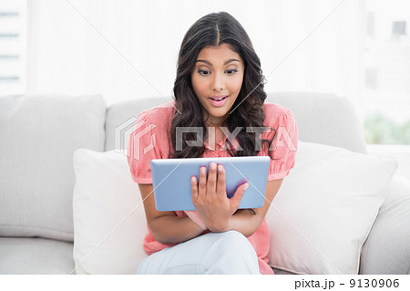 Excited cute brunette sitting on couch using tablet Excited cute brunette sitting on couch using tablet 9130906