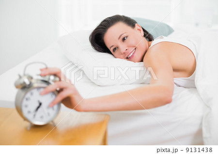 Smiling woman in bed extending hand to alarm clock 9131438