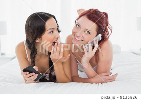 Shocked woman looking at a cheerful friend while on call in bed 9135826