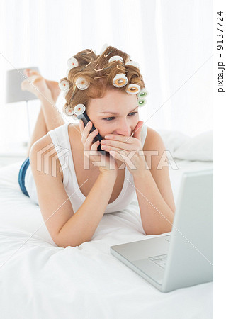 Shocked woman in hair curlers using cellphone and laptop in bed 9137724