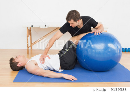 Physical therapist assisting young man with yoga ball 9139583