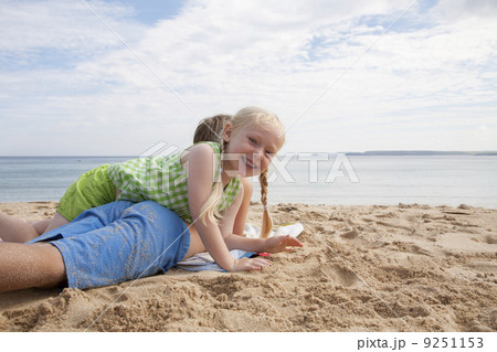 A boy lying on his front on the sand looking out to sea. His sister lying on top of him.   9251153