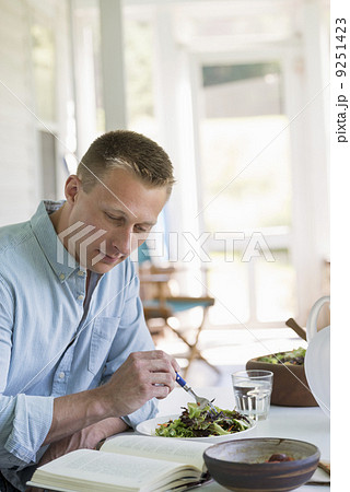 A man seated at a cafe table eating a salad and reading a book. A man seated at a cafe table eating a salad and reading a book. 9251423
