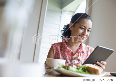 A young woman reading from the screen of a digital tablet, seated at a table. Coffee and a sandwich.  9251770