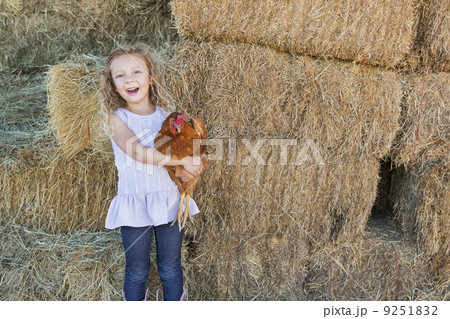 A young girl standing in a hay barn holding a chicken in her arms.  9251832