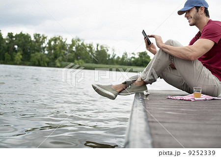 A man seated on a jetty by a lake, using a digital tablet. A man seated on a jetty by a lake, using a digital tablet. 9252339