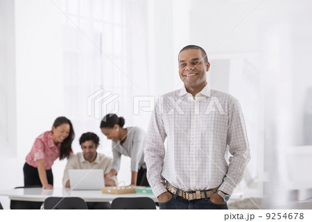 Business. A Man Standing With His Hands In His Pockets, And Three People Leaning Over A Desk Behind Him. Business. A Man Standing With His Hands In His Pockets, And Three People Leaning Over A Desk Behind Him. 9254678