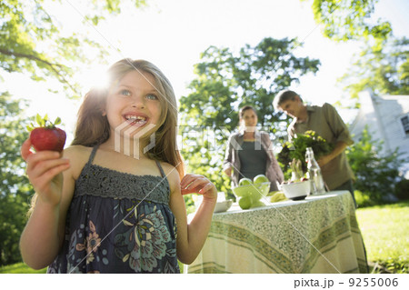 On The Farm. Children And Adults Together. A Young Girl Holding A Large Fresh Organically Produced Strawberry Fruit. Two Adults Beside A Round Table. On The Farm. Children And Adults Together. A Young Girl Holding A Large Fresh Organically Produced Strawberry Fruit. Two Adults Beside A Round Table. 9255006