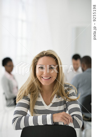 Office Interior. A Woman In A Striped Shirt Seated Astride A Chair Looking Away From A Group Seated At A Table. 9255198