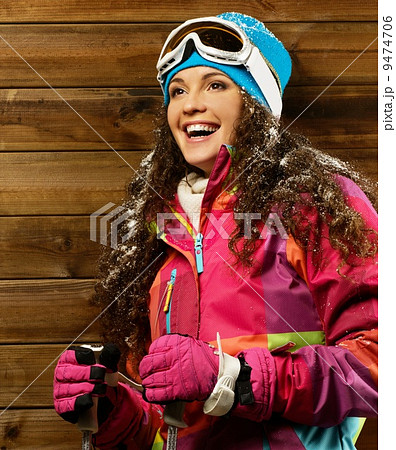Smiling woman with ski poles standing against wooden house wall Smiling woman with ski poles standing against wooden house wall 9474706