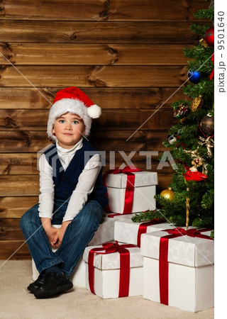 LIttle boy in Santa hat with gift box under christmas tree in wooden house interior LIttle boy in Santa hat with gift box under christmas tree in wooden house interior 9501640