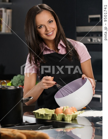 Young woman in apron whisking eggs for a dough on a modern kitchen Young woman in apron whisking eggs for a dough on a modern kitchen 9501741