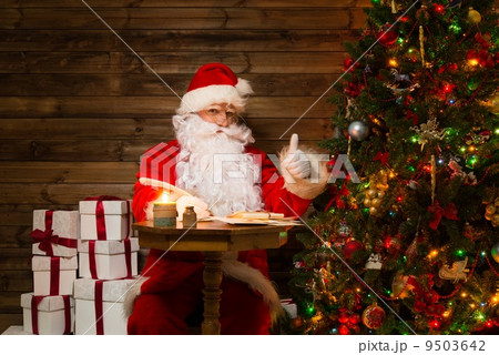 Santa Claus in wooden home interior sitting behind table and writing letters with quill pen 9503642