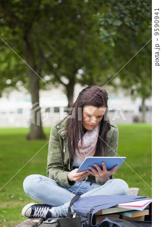 Focused brunette student using tablet sitting on bench Focused brunette student using tablet sitting on bench 9590941