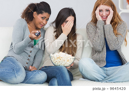 Scared friends with remote control and popcorn bowl on sofa 9645420