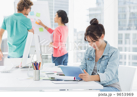 Casual woman using digital tablet with colleagues behind in offi Casual woman using digital tablet with colleagues behind in offi 9653969